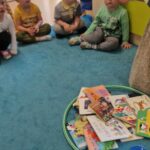 Dzieci siedzą w rzędzie na turkusowym dywanie i patrzą na opiekunkę, która pokazuje im zniszczoną książeczkę. Przed dziećmi na dywanie leżą dwa zielone hula-hop. W jednym znajdują się książeczki w dobrym stanie a w drugim zniszczone.