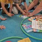 Dzieci siedzą na turkusowym dywanie i pomagają opiekunce naprawić zniszczoną książeczkę. Przed dziećmi na dywanie leżą dwa zielone hula-hop. W jednym hula-hop znajdują się książeczki w dobrym stanie a w drugim zniszczone.
