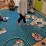Dziewczynka stoi i patrzy na kolorowe książeczki rozłożone na dywanie, obok leżą dwa zielone hula-hop. W jednym hula-hop znajdują się książeczki w dobrym stanie a w drugim zniszczone książeczki.