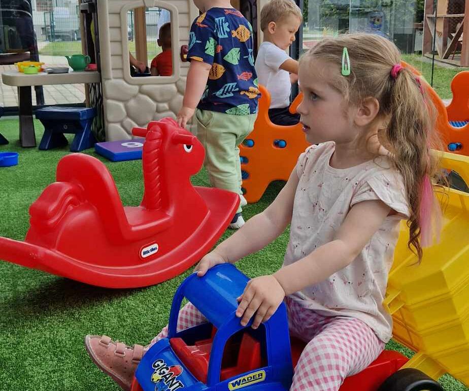 Dziewczynka siedzi na kolorowym zabawkowym samochodziku. W tle widać altanę i bawiące się w niej dzieci. W altanie znajduje się duży szary domek i kolorowe zabawki.