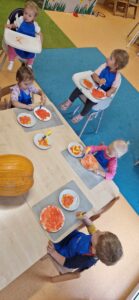 Ubrane w granatowe fartuszki dzieci siedzą przy stole i malują pomarańczową farbą papierowe talerzyki. Na środku stołu leży duża dynia.