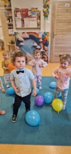 Chłopiec wraz z koleżankami tańczy na turkusowym dywanie. W tle widać urodzinową dekorację.