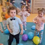 Chłopiec wraz z koleżankami tańczy na turkusowym dywanie. W tle widać urodzinową dekorację.