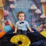 Chłopiec siedzi na ciemno granatowym materiale, w tle widać urodzinową dekorację.