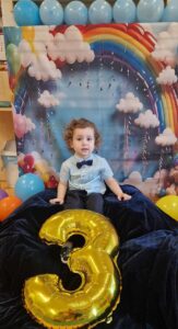 Chłopiec siedzi na ciemno granatowym materiale, w tle widać urodzinową dekorację.