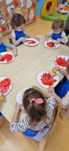 Dzieci siedzą przy stoliku i malują czerwoną farbą białe papierowe talerzyki.