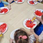 Dzieci siedzą przy stoliku i malują czerwoną farbą białe papierowe talerzyki.