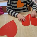 Chłopiec siedzi przy stole i łączy w pary papierowe czerwone serduszka.