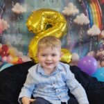 Chłopiec siedzi na granatowym materiale i uśmiecha się do zdjęcia. W tle widać urodzinową dekoracje.