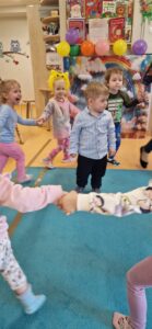 Dzieci trzymają się za rączki i tańczą w kółeczku. W środku stoi solenizant.
