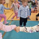 Dzieci trzymają się za rączki i tańczą w kółeczku. W środku stoi solenizant.