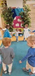 Dzieci ubrane na kolor niebieski trzymają się za ręce i tańczą w kole. W tyle widać wioskę smerfów.