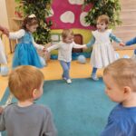 Dzieci ubrane na kolor niebieski trzymają się za ręce i tańczą w kole. W tyle widać wioskę smerfów.