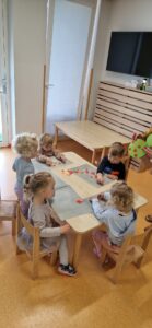 Dzieci siedzą przy stoliku i naklejają kolorowe kółeczka tworząc z nich drzewo.