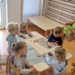 Dzieci siedzą przy stoliku i naklejają kolorowe kółeczka tworząc z nich drzewo.