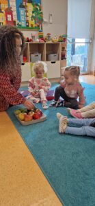 Dzieci siedzą na turkusowym dywanie i oglądają owoce jabłko, gruszkę i śliwki. W tle widać sale zabaw.