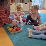Dzieci siedzą na turkusowym dywanie i oglądają owoce jabłko, gruszkę i śliwki. W tle widać sale zabaw.