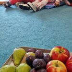 Dzieci siedzą na podusiach i oglądają owoce z sadu.