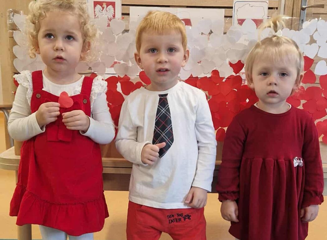 Dzieci stoją koło siebie ubrane na biało- czerwono. W tle widać flagę biało- czerwoną