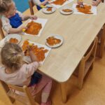 Dzieci siedzą przy stoliku i malują brązową farbą na białej kartce odrysowane rączki.