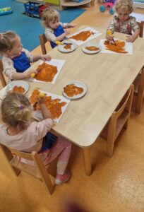 Dzieci siedzą przy stoliku i malują brązową farbą na białej kartce odrysowane rączki.