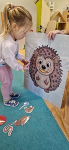 Dziecko stoi przed tekturowym jeżem i wkłada do wyciętej buzi tekturowe jedzenie. W tle widać sale zabaw.