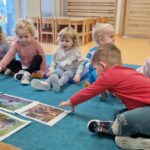 Dzieci siedzą na turkusowym dywanie i oglądają fotografię z jeżami. W tyle widać sale zabaw.