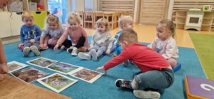 Dzieci siedzą na turkusowym dywanie i oglądają fotografię z jeżami. W tyle widać sale zabaw.